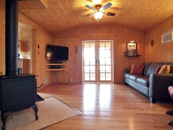 A wooden interior with a black stove, a television on a shelf, a sofa, and a ceiling fan.