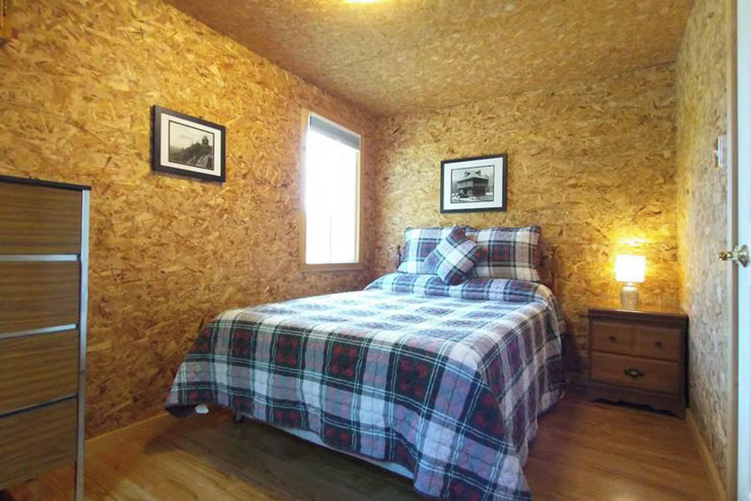 Un dormitorio con pared de madera, una cama con colcha escocesa, una mesita de noche con lámpara y una ventana.