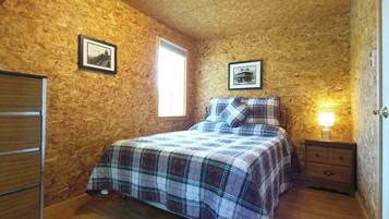 Un dormitorio con pared de madera, una cama con colcha escocesa, una mesita de noche con lámpara y una ventana.