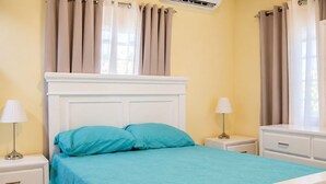 Ein Bett mit weißem Kopfteil, zwei Kissen und einer petrolfarbenen Bettdecke in einem Zimmer mit gelben Wänden, einem fenster mit Vorhängen und einem Nachttisch mit einer Lampe.