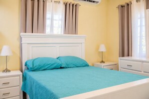 A bed with a white headboard, two pillows, and a teal bedspread in a room with yellow walls, a window with curtains, and a bedside table with a lamp. - Perfect 7-bedroom Village with AC in awesome Higüey location (Higüey)