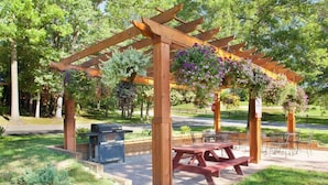 Una pèrgola de fusta amb cistells de flors penjants, una barbacoa i taules de pícnic en un parc.