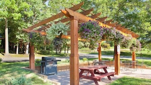 Una pérgola de madera con macetas colgantes de flores, una parrilla para barbacoa y mesas de picnic en un parque.