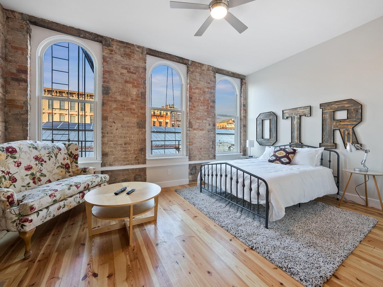 A bedroom with a large bed, a sofa, a small table, and a view of buildings through large windows.