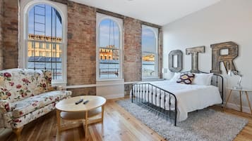A bedroom with a large bed, a sofa, a small table, and a view of buildings through large windows.