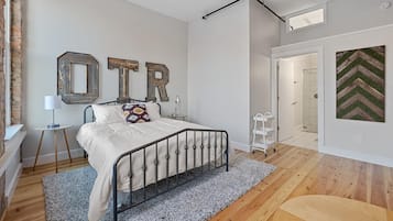 A bedroom with a bed, a small table, a lamp, a wall-mounted mirror, and a door leading to a bathroom.