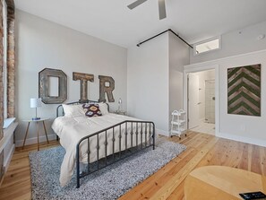 A bedroom with a bed, a small table, a lamp, a wall-mounted mirror, and a door leading to a bathroom. - Findlay Suites Boutique Apart-Hotel (Cincinnati)