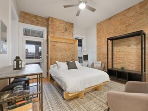A bedroom with a bed, a desk, a chair, a TV, and a brick wall. - Findlay Suites Boutique Apart-Hotel (Cincinnati)