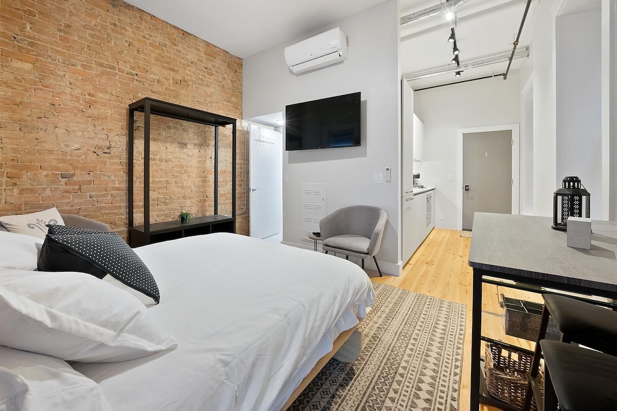 A modern bedroom with a brick wall, a bed with white bedding, a desk with a chair, and a television.