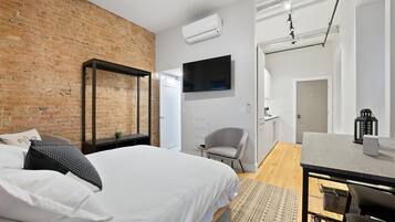 A modern bedroom with a brick wall, a bed with white bedding, a desk with a chair, and a television.