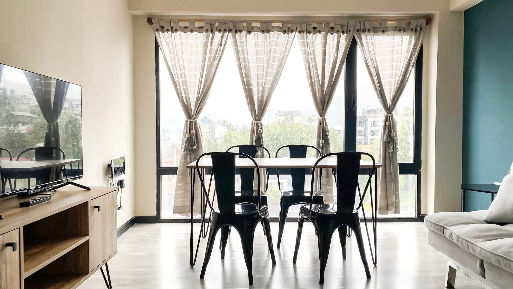 A modern living room with a dining area, large windows with curtains, a flat-screen TV, and a sofa.
