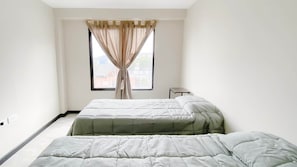 Ein Zimmer mit zwei Betten, einem Fenster mit Vorhängen und einem kleinen Tisch.