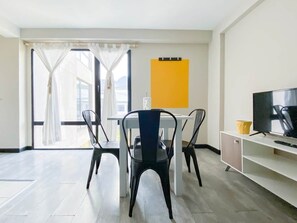 Ein modernes Wohnzimmer mit einem Esstisch, vier Stühlen, einem Flachbildfernseher und einem großen Fenster mit Vorhängen.