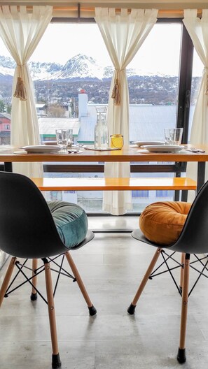 Ein Essbereich mit einem Tisch für zwei Personen, Stühle mit Kissen, Blick auf eine gebirgige Landschaft und ein Fenster mit Vorhängen.