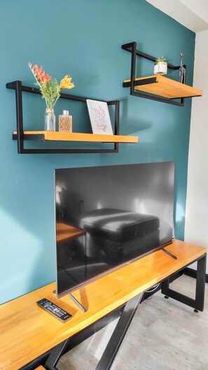 Ein Zimmer mit türkisfarbenen Wänden, einem Holztisch mit Fernseher und zwei schwarzen Metallregalen mit Dekorationselementen.