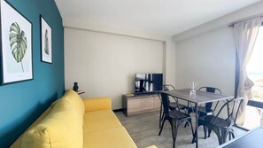 Un espace de vie moderne avec un canapé jaune, une table à manger avec des chaises, un téléviseur à écran plat et des reproductions botaniques encadrées accrochées au mur.