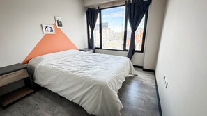 A bedroom with a bed, a nightstand, a window with curtains, and a view of buildings outside. - Amanecer del Fuego VI (Ushuaia)