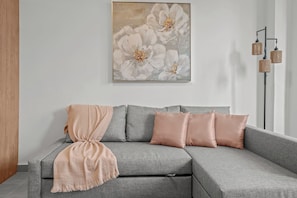 Ein graues Ecksofa mit einer pfirsichfarbenen Decke und passenden Kissen, ein großes Blumenbild an der Wand und eine stehende Stehlampe mit drei Lampenschirmen.