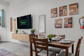 Ein moderner Essbereich mit einem Holztisch, gedeckt für vier Personen, einem an der Wand montierten Flachbildfernseher, der 'hulu' zeigt, und abstrakten Kunstwerken an den Wänden.
