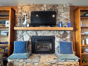 A stone fireplace with a television above it, flanked by two blue cushioned benches. - Juniper Beach Cabin on Camano Island (Camano Island)