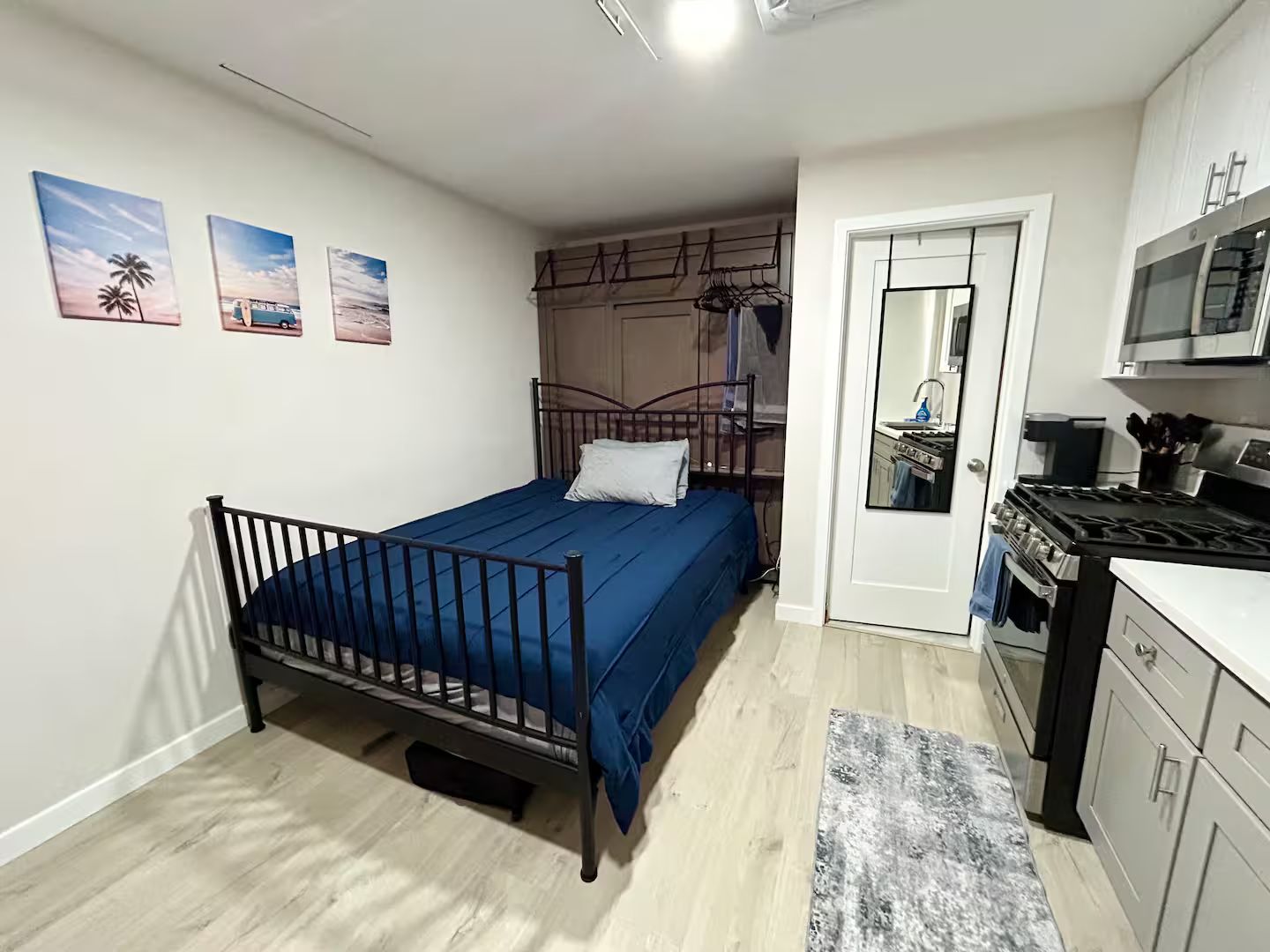 A bedroom with a bed, a wardrobe, a kitchen area with a stove and microwave, and a door leading to another room.