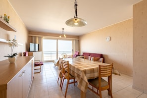 A dining area with a table set for four, wooden chairs, a sofa, and a television on a cabinet. - Apartment in Blankenberge With Sea Views (Blankenberge)