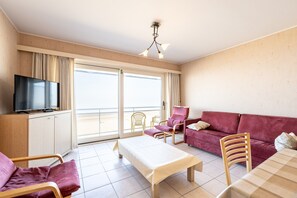 A room with a television, a sofa, a chair, a table, and a balcony with a view. - Apartment in Blankenberge With Sea Views (Blankenberge)