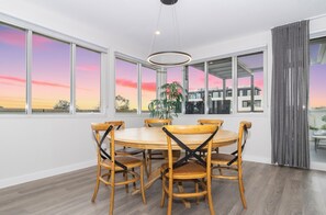 A dining area with a round table and six chairs, large windows with a view of a sunset, and a hanging light fixture. - Corner Penthouse with City Views in Armadale (Armadale)
