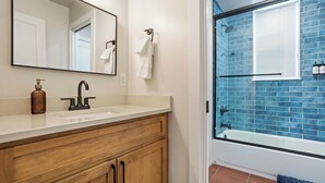 Un baño con tocador de madera, un espejo grande, un lavamanos blanco y un área de ducha con mampara con azulejos azules.