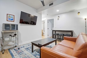 Una sala moderna con un sofá de cuero, un televisor de pantalla plana montado en la pared, una zona de cocina con microondas y una mesa de comedor con sillas.