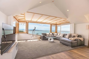 A modern living room with a large sectional sofa, a flat-screen TV, a telescope, and a view of the sea through large windows.