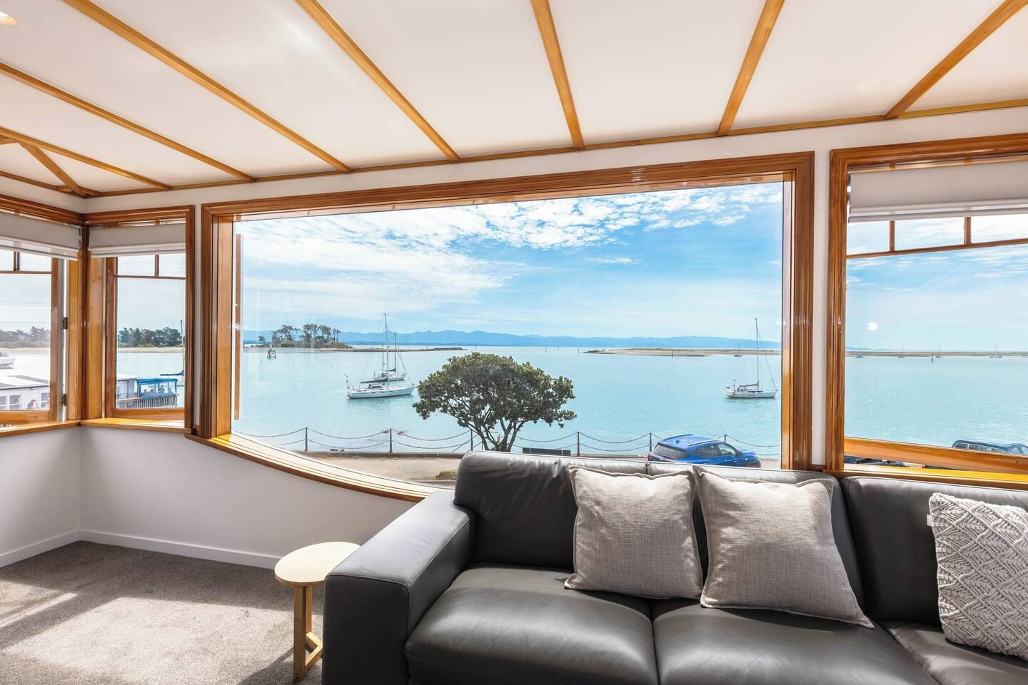A room with a black leather sofa, a small round table, and large windows offering a view of a marina with boats and a tree.