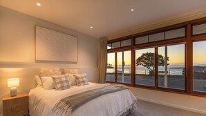 Ein Schlafzimmer mit einem großen Bett, einem Nachttisch mit Lampe, Blick auf das Meer durch große Fenster und einem Gemälde an der Wand.
