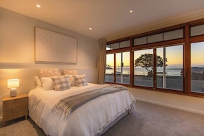 Un dormitorio con una cama grande, una mesita de noche con lámpara, vista al mar a través de amplios ventanales y un cuadro en la pared.