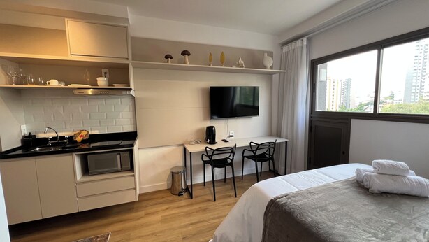 A compact hotel room with a kitchenette, a bed with a neatly folded towel, a TV mounted on the wall, and a window offering a city view. - Vrbo Property (São Paulo)