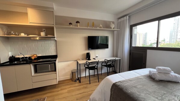 Kompakti hotellihuone, jossa on pieni keittiönurkkaus, siististi taitettu pyyhe sängyllä, seinälle asennettu televisio ja ikkuna, josta avautuu näkymä kaupunkiin.