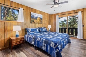 Un dormitorio con piso de madera, una cama con colcha azul y blanca a cuadros, dos mesitas de noche con lámparas, un ventilador de techo y un ventanal con cortinas.