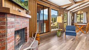 A room with a brick fireplace, wooden flooring, a dining area with a table and chairs, and large windows offering a view of the outdoors. - Machins House Newly Remodled 4 bdrm backs up against RMNP (Estes Park)