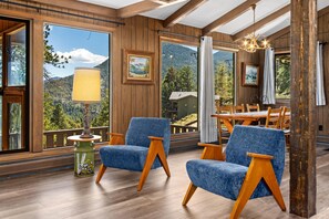 Ein Zimmer mit Holzwänden, einem Essbereich mit Stühlen und einem Tisch sowie großen Fenstern mit Blick auf die Berge.