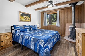 A bedroom with a large bed, wooden headboard, a painting, a window with curtains, a ceiling fan, and a wood-burning stove. - Machins House Newly Remodled 4 bdrm backs up against RMNP (Estes Park)