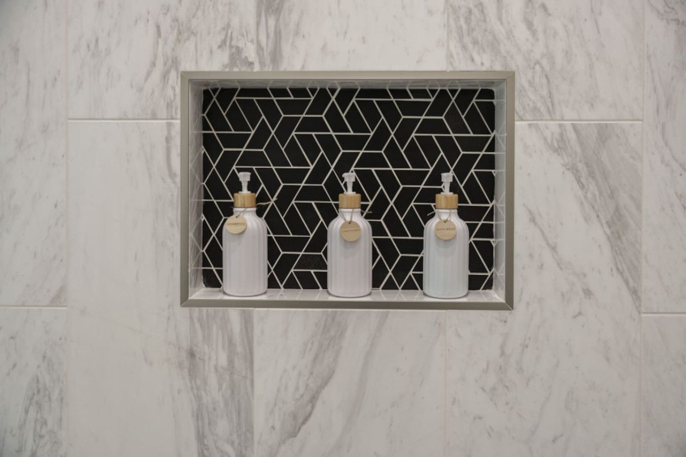 Un baño con pared de mármol y un estante de diseño geométrico en negro que sostiene tres botellas blancas con tapas de madera.