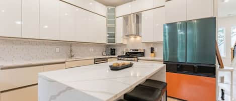 Una cocina moderna con armarios blancos, una isla de mármol, taburetes negros y un refrigerador grande.