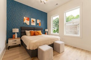 Een slaapkamer met een blauw behang, een bed met een grijze hoofdbord, twee nachtkastjes met lampen, en een raam met uitzicht op groen.