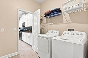 A laundry room with two front-loading washers and dryers, a drying rack with folded towels, and a kitchen area visible through an open door. - Perfect Family Escape, Pool, Themed Rooms (Kissimmee)