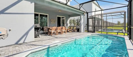 An indoor swimming pool with a sunlit patio area, a barbecue grill, and a view of the surrounding landscape.