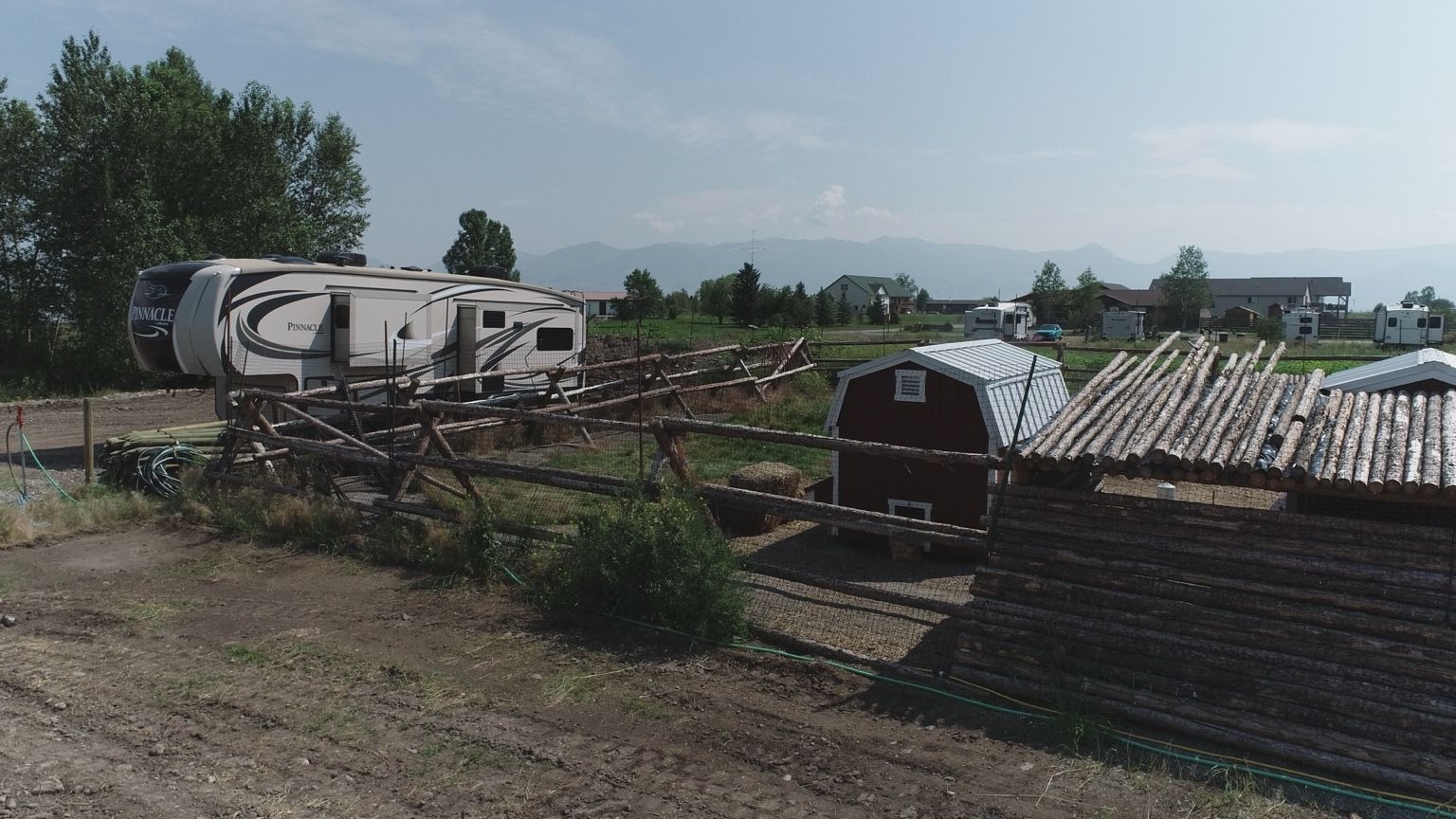 A caravan parked in a rural setting with a wooden structure and a red barn in the background.