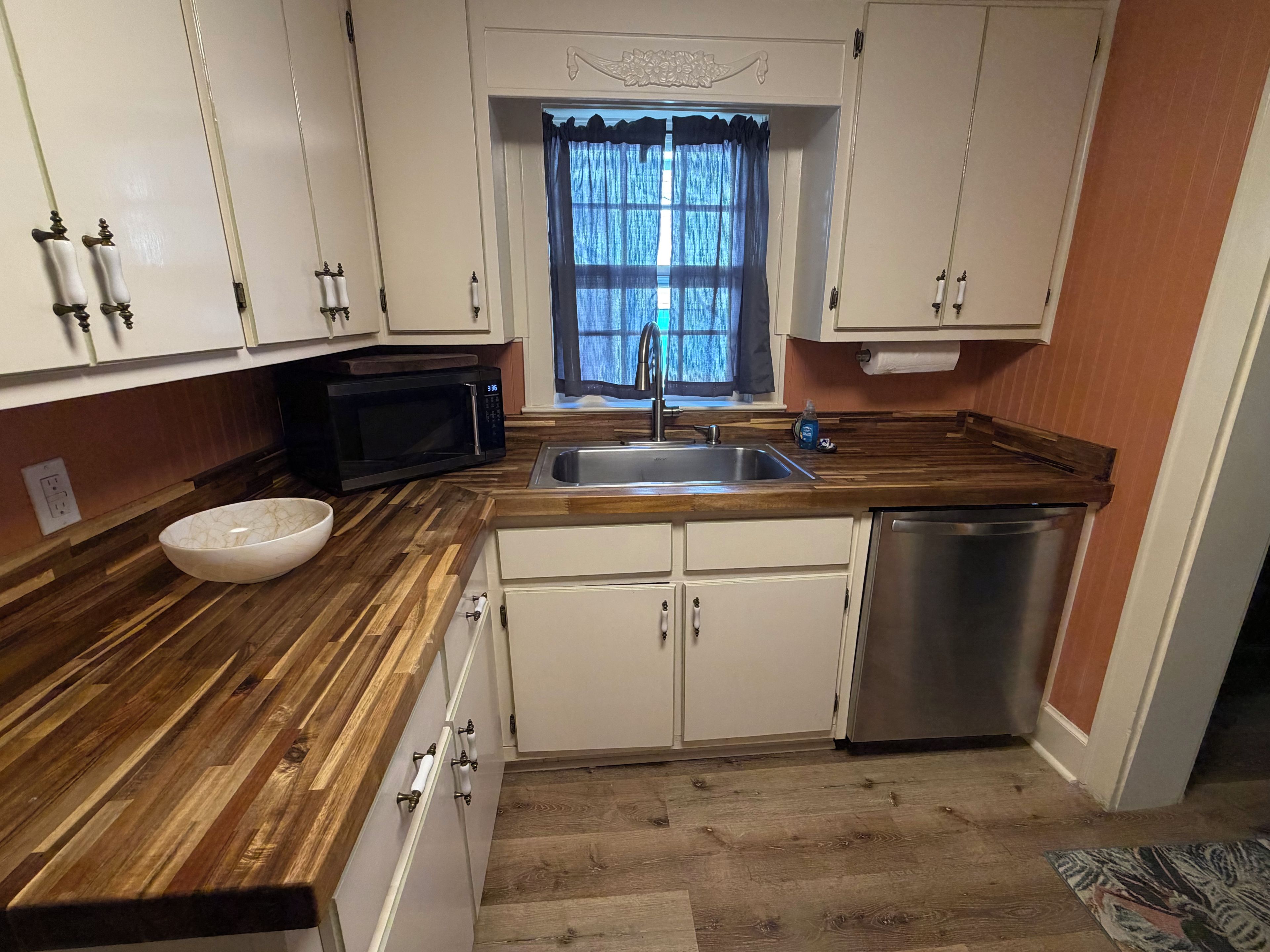 A kitchen with a wooden countertop, white cabinets, a stainless steel sink, a microwave, and a dishwasher.