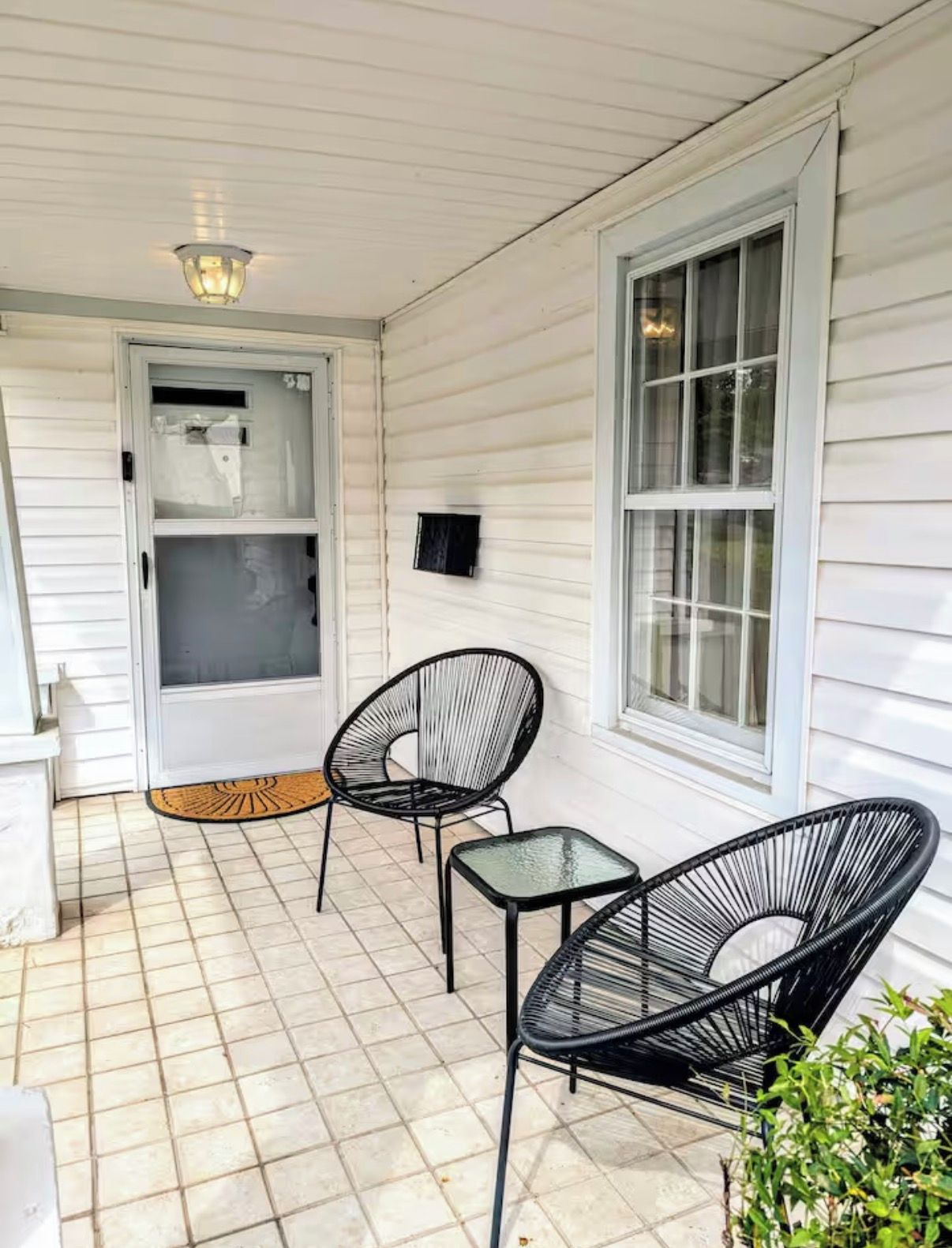 Een overdekte veranda met twee zwarte metalen stoelen en een kleine tafel, een deur met hor en een wandlamp.