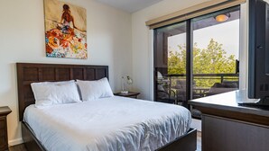 Une chambre avec un grand lit, une table de chevet, un bureau, une chaise et un balcon donnant sur les arbres.