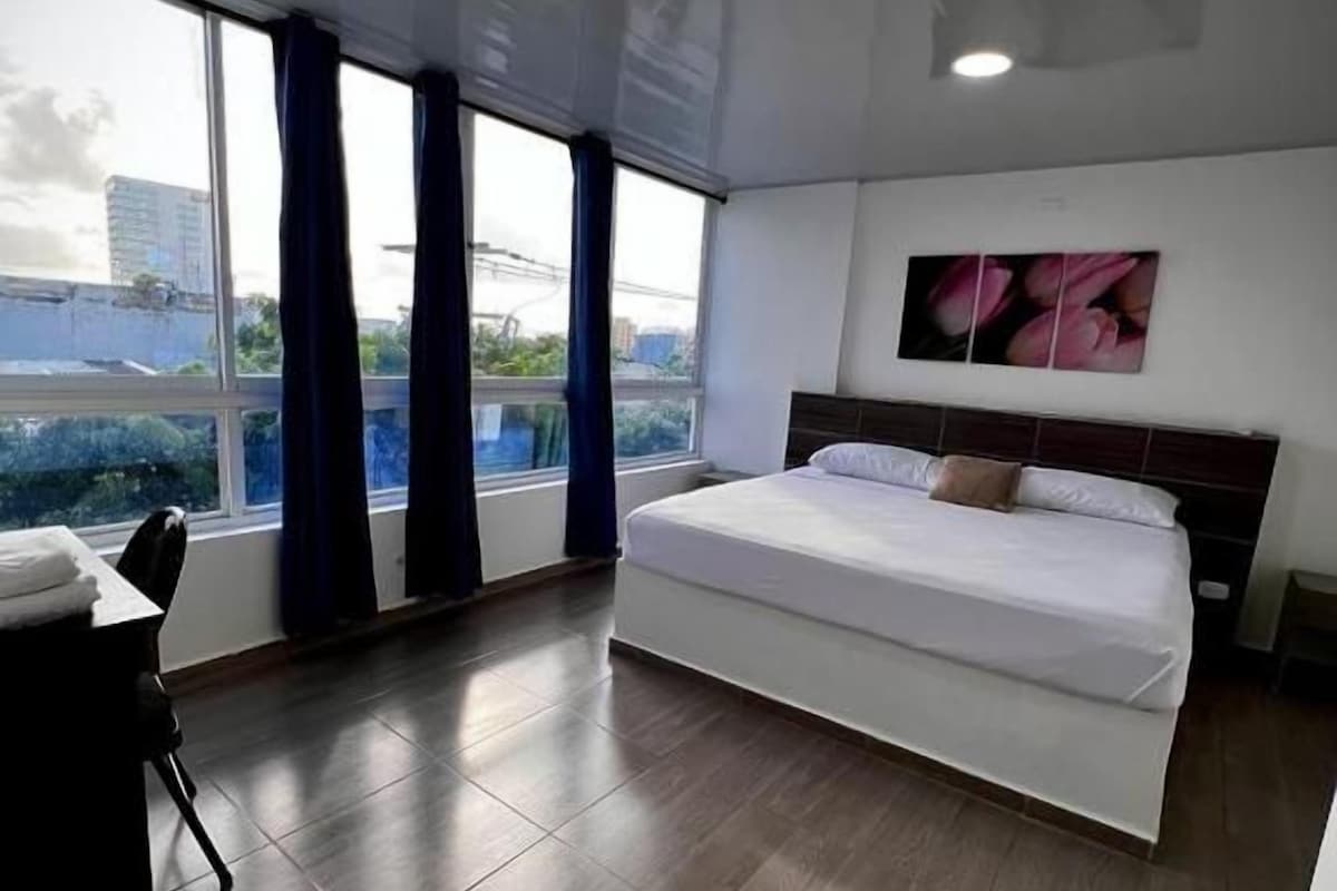 A modern hotel room with a large bed, a desk, and a view through a window.